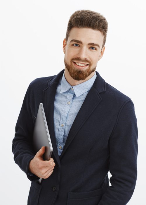 Stylish confident and sexy male entrepreneur with blue eyes and beard standing self-assured in formal suit holding laptop in hand gazing pleased and assured at camera, being ambitious and successful. Technology, finance and people concept