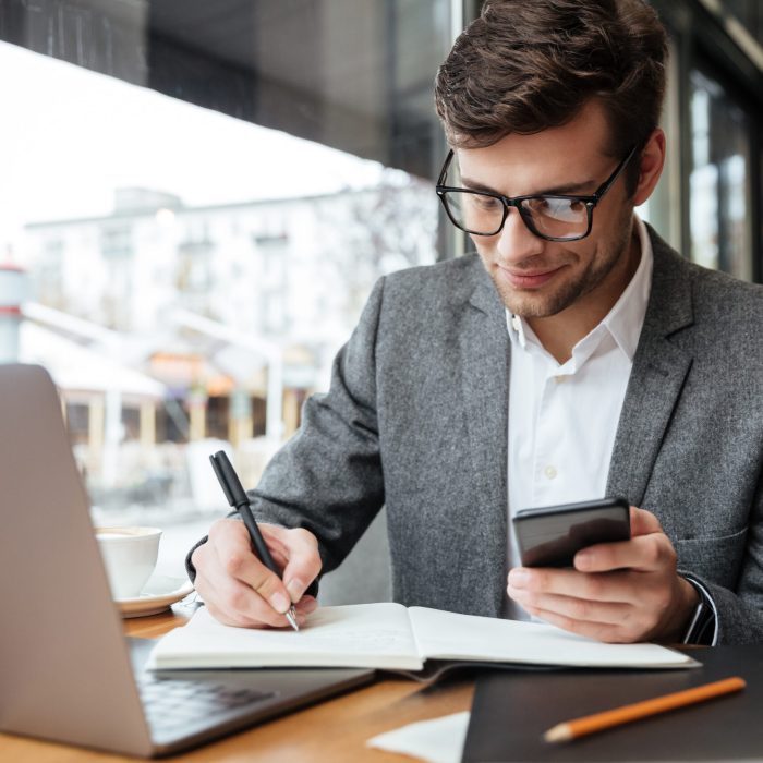 Smiling business man in eyeglasses sitting by the table in cafe with laptop computer while using smartphone and writing something